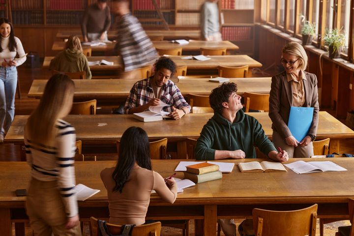 Blid, kvinnelig professor snakker med mannlig student i biblioteket med flere studenter rundt