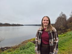 Kayla Buhler står smilende ved en elvebredd, iført en rutet jakke, med skog og vann i bakgrunnen under en grå himmel.