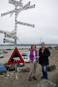 Prorektor for forskning og utvikling ved UiT, Camilla Brekke (t.v.) på flyplassen i Longyearbyen, sammen med Sandrine Lacombe, som er professor og viserektor ved Paris Saclay Université og leder av EUGLOH. De står ved siden av et skilt med isbjørn og avstand til mange forskjellige byer.
