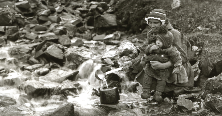 Eldre sort-hvitt fotografi av en samisk mor som sitter med et lite barn ved en bekk og øser vann oppi en kaffekjele.