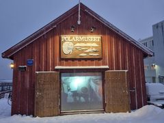 Bildet viser inngangen til Polarmuseet i Tromsø, en rød trebygning dekket med snø. En stor plakat med teksten "POLARMUSEET" vises over døren.