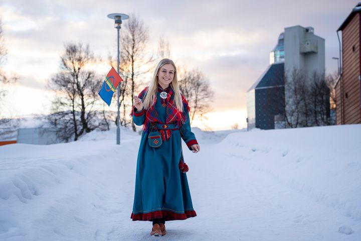 Kvinne kledd i kofte går med samisk flagg på et vinterkledd universitetsområde.