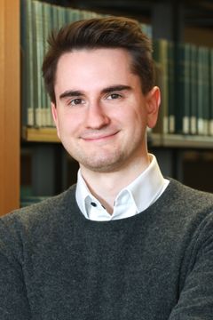En smilende person med mørkt hår og grå genser står foran en bokhylle.