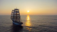 A tall ship with full sails on the ocean at sunset.