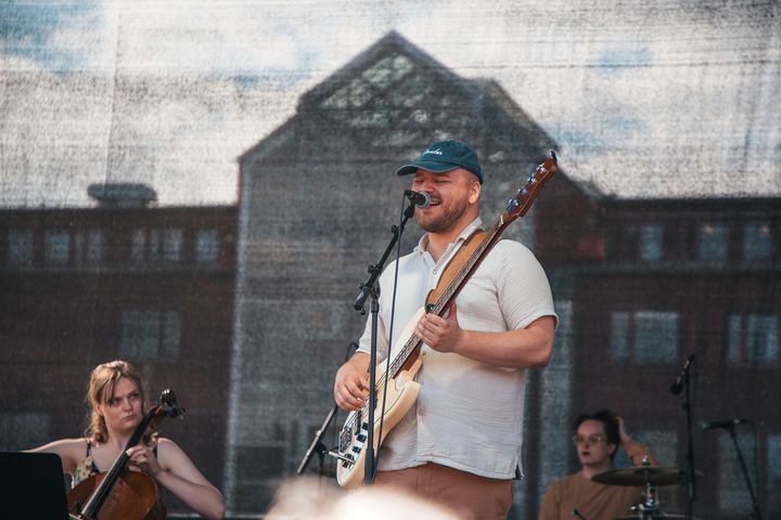 Mann spiller el-gitar og synger med innlevelse foran universitetsbygning.