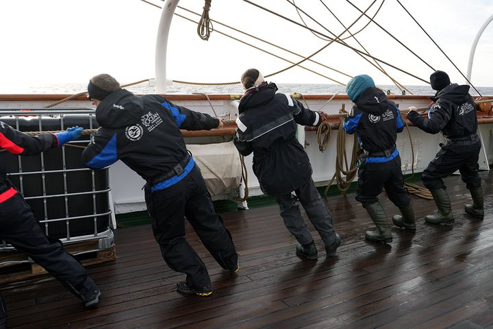 Personer i seiledresser drar i tau på dekk under en seiling.
