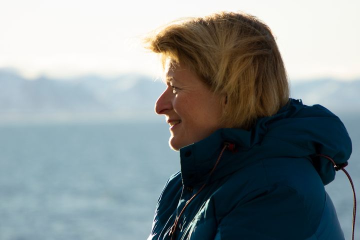 Person med blondt hår i profil, kledd i blå jakke, ser utover havet med fjell i bakgrunnen.