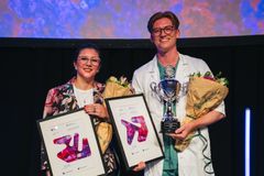 To personer holder diplomer, blomster og en pokal på en scene.