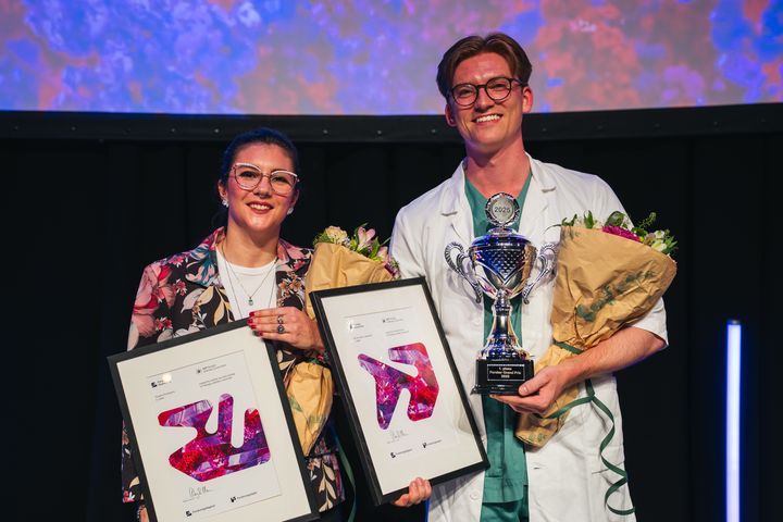To personer holder diplomer, blomster og en pokal på en scene.