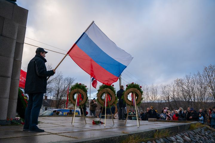 En person holde det russiske flagget i bakgrunnen, mens en mann holder en tale foran en statue