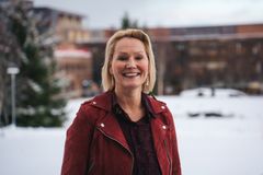 dame med blondt hår som smiler og står ute i et vinterlandskap med noen bygg i bakgrunnen