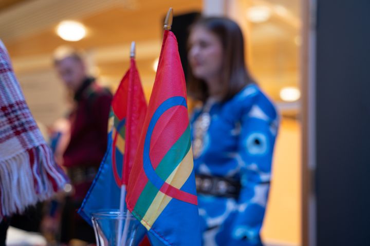 To samiske flagg står plassert i forgrunnen, med personer i tradisjonelle klær uskarpt i bakgrunnen.