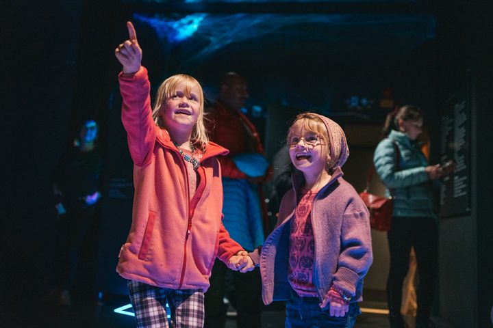 To barn smiler og peker oppover, mens de holder hender i et dunkelt opplyst rom.