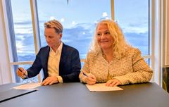 To personer signerer en samarbeidsavtale ved et bord med utsikt over et vinterlandskap utenfor vinduet.