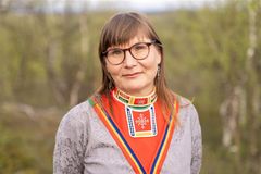 En kvinne med langt hår og briller, ikledd tradisjonell samisk drakt, står utendørs med trær i bakgrunnen.