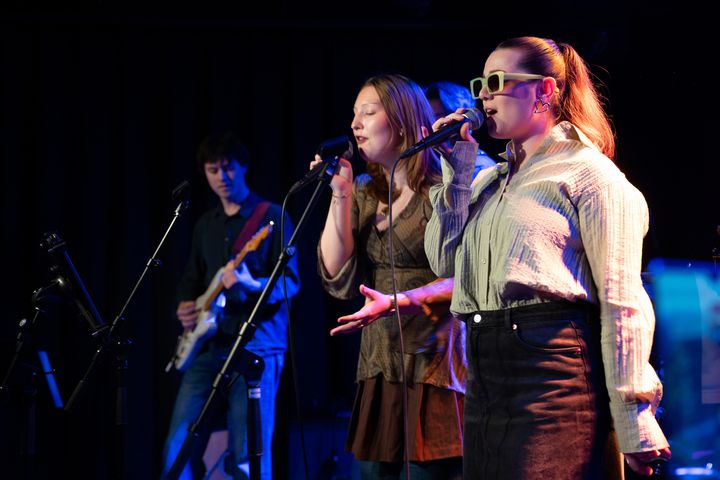 To kvinner står på scene og synger, i bakgrunnen en mann som spiller gitar.