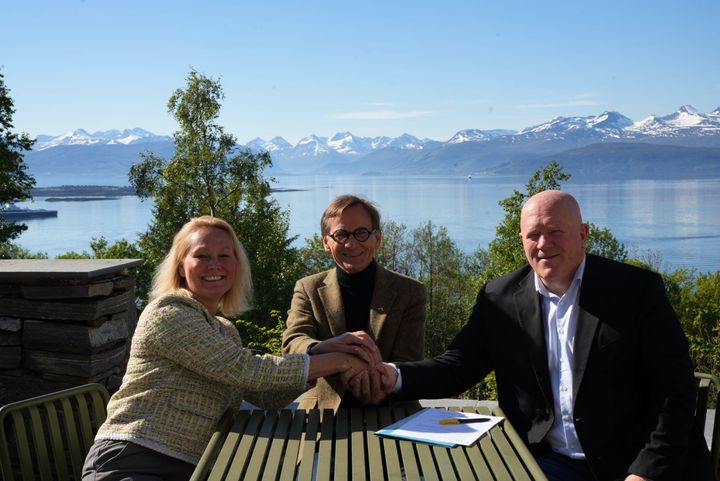 En kvinne og to menn signerer avtale foran fjord og fjell. Vår og blå himmel.
