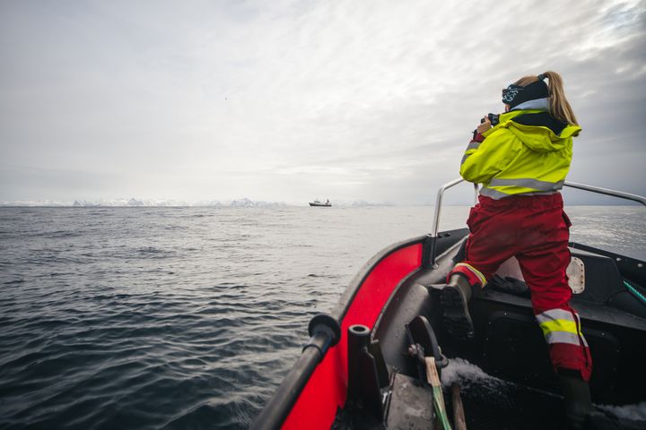 En person i rød og gul beskyttelsesdrakt står på en båt og ser ut over et åpent hav med en båt i horisonten.