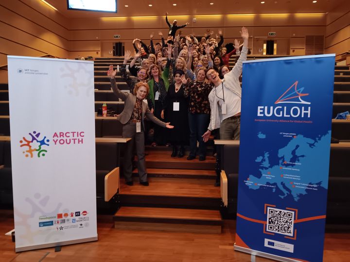 Deltakere smiler og løfter armene under EUGLOH-konferansen om sosialt arbeid på UiT i Tromsø, 2025. To bannere vises foran gruppen.