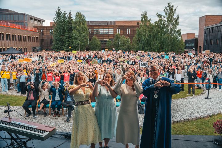 Et knippe rektorer og politikere viser hjertetegn med hendene på en scene med masse glade, forventningsfulle studenter i bakgrunnen.