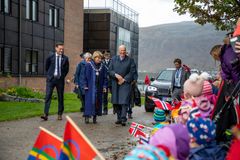 En gruppe mennesker går forbi barn som holder norske flagg. En bil og fjell er i bakgrunnen.