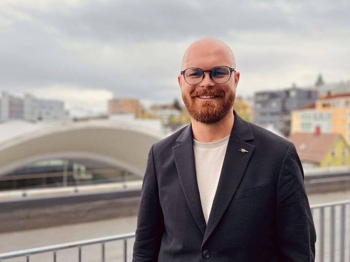 Portrett av en person som smiler, har skjegg, briller og står utendørs ved et rekkverk. I bakgrunnen er moderne bygninger og en overskyet himmel.