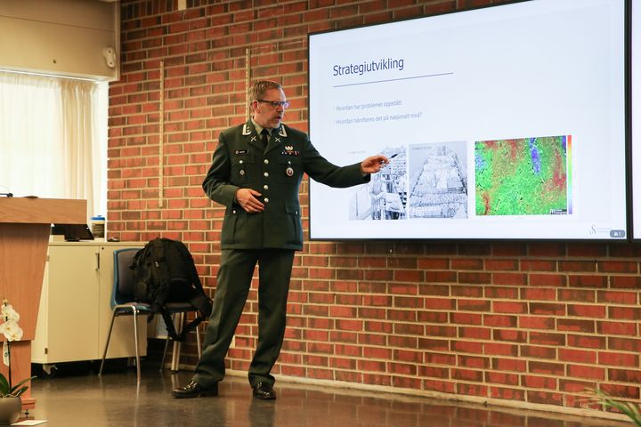 Uniformert offiser står på podium og forklarer en presentasjon med bilder og overskriften "Strategiutvikling".