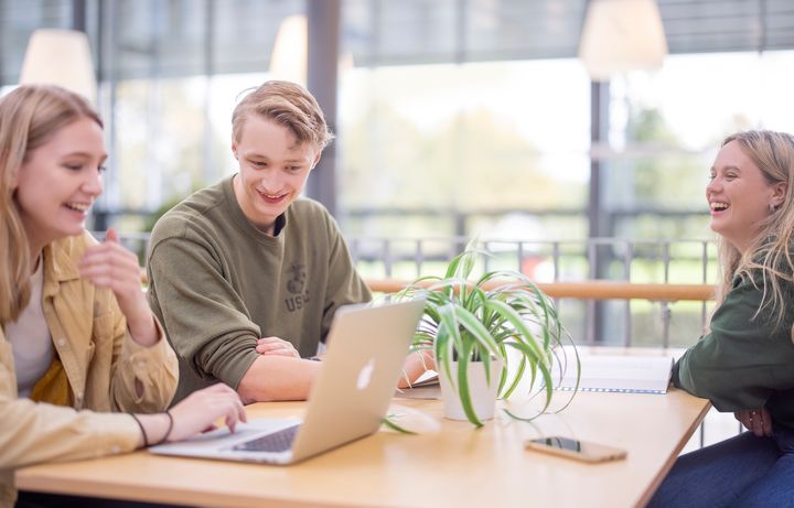 Tre studenter sitter rundt et bord i et lyst lokale med en pc på bordet foran seg