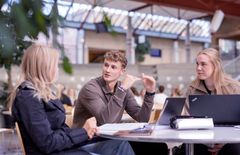 Tre studenter samarbeider diskuterer rundt et bord i et atrium