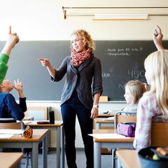 kvinne står i et klasserom foran en tavle. Hun smiler og i rommet er det mange barn. Hun holder en hånd opp for å peke og tilsynelatende gi ordet til en av elevene i rommet.