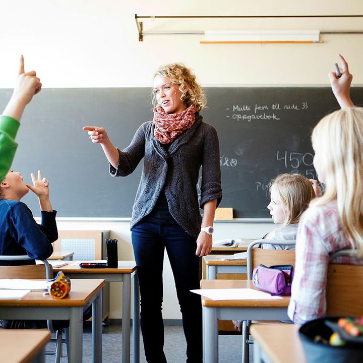 kvinne står i et klasserom foran en tavle. Hun smiler og i rommet er det mange barn. Hun holder en hånd opp for å peke og tilsynelatende gi ordet til en av elevene i rommet.