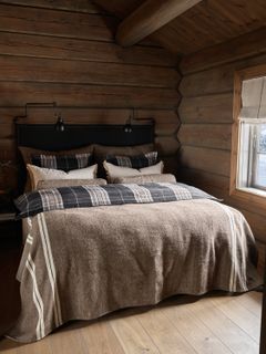 Et koselig soverom med trevegger, en dobbeltseng med rutete sengetøy, og en vindu til høyre.