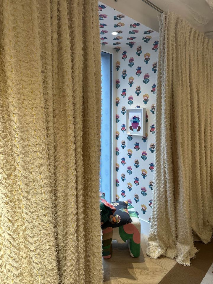 Tykk%20beige%20gardinrammer%20inn%20en%20liten%20stue%20med%20blomstrete%20tapet%20og%20fargerik%20stol.