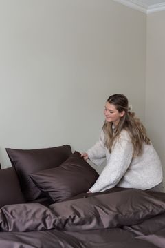 En kvinne i beige strikkegenser ordner mørkebrune sengeklær og puter på en seng.