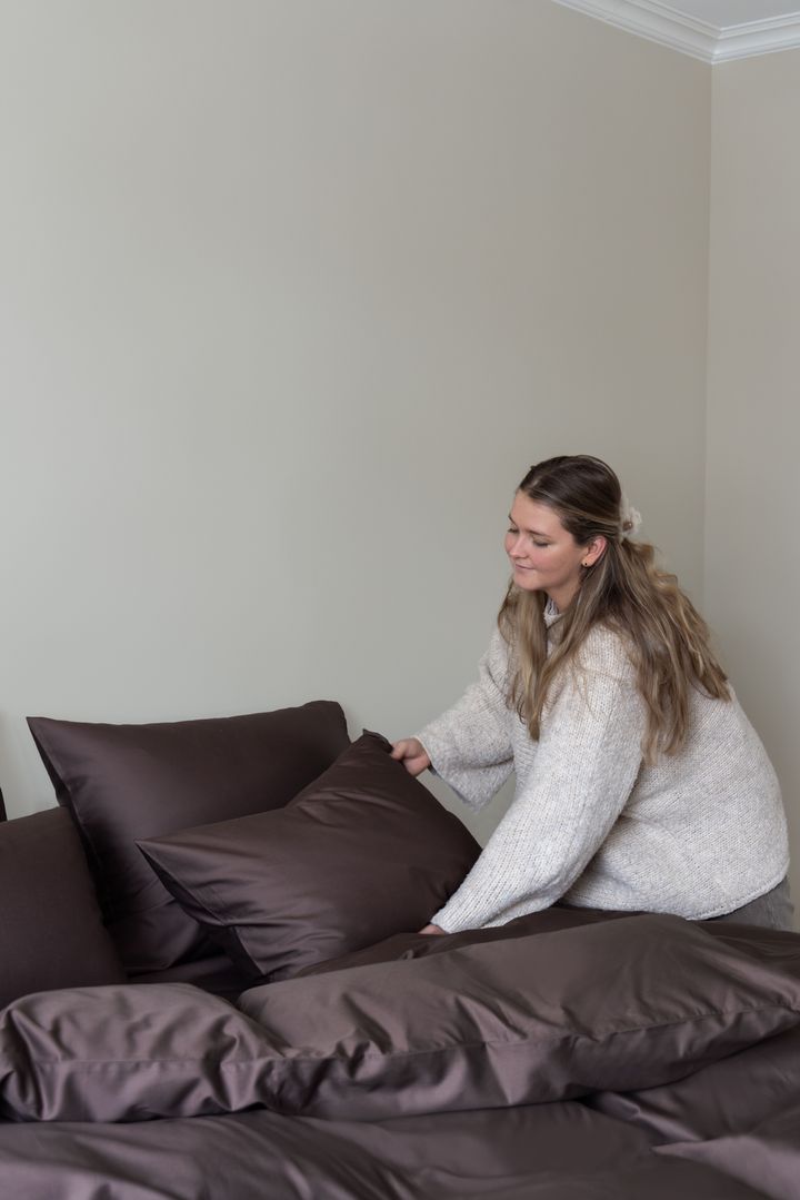 En%20kvinne%20i%20beige%20strikkegenser%20ordner%20m%F8rkebrune%20sengekl%E6r%20og%20puter%20p%E5%20en%20seng.
