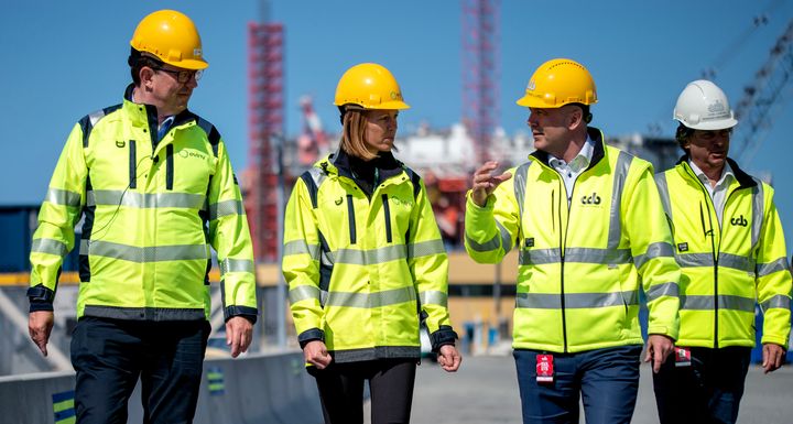 Fire personer i gul vernejakke og hjelm som går inne på et industriområde med en plattform i bakgrunnen.