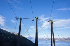 To personer jobber på toppen av høye kraftlinjestolper med en fjord og fjell i bakgrunnen.