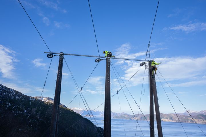 To%20personer%20jobber%20p%E5%20toppen%20av%20h%F8ye%20kraftlinjestolper%20med%20en%20fjord%20og%20fjell%20i%20bakgrunnen.