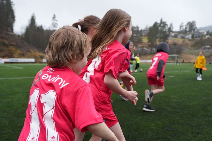 Barn i røde Eviny-drakter spiller fotball på en gressbane.