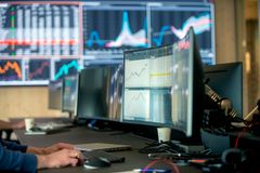 En person som jobber ved en dataskjerm med grafer og tall, med flere skjermer i bakgrunnen som viser ulike data og diagrammer.