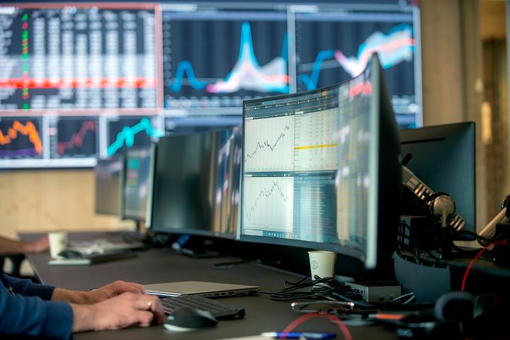 En person som jobber ved en dataskjerm med grafer og tall, med flere skjermer i bakgrunnen som viser ulike data og diagrammer.