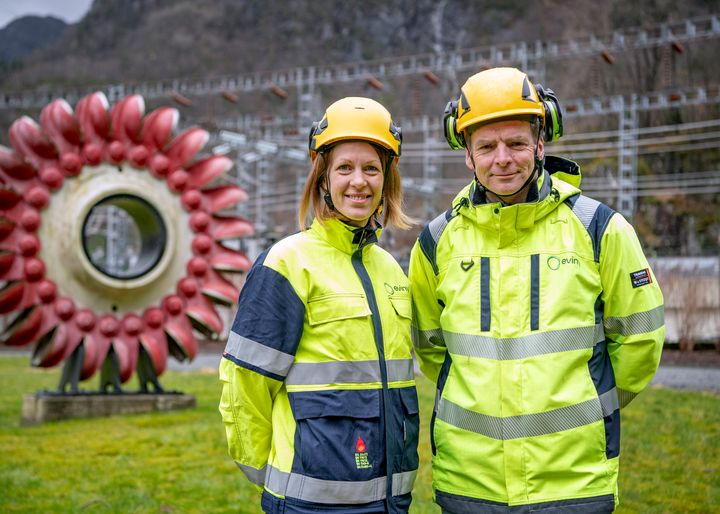 To personer i gult vernetøy og hjelm står foran et indstrianlegg utendørs.