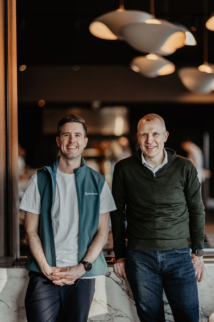 To personer som står sammen og smiler, innendørs.