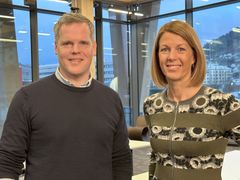 To personer smiler foran store vinduer med utsikt.