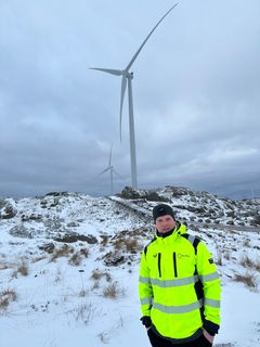 Mann i gul jakke står foran vindturbiner i et snødekt landskap.