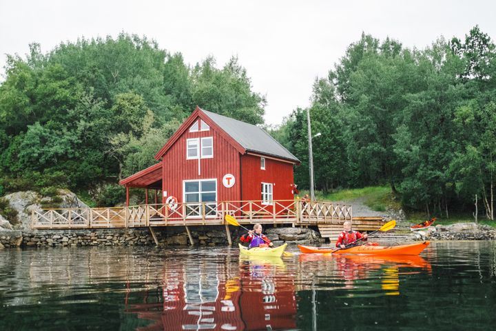 To personer padler i kajakker foran et rødt hus ved vannet.