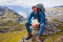 En mann med turklær og ryggsekk sitter på en stein med fjellandskap i bakgrunnen.