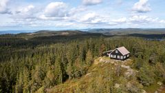 Liten svart hytte på en høyde omgitt av skog og fjell under en delvis skyet himmel.