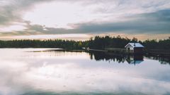 Rolig innsjø med et lite trehus ved vannkanten og skog i bakgrunnen. Skyet himmel reflekteres i det stille vannet.