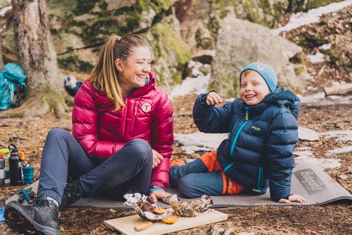 En kvinne og et barn smiler mens de sitter på bakken i en skog og spiser mat.